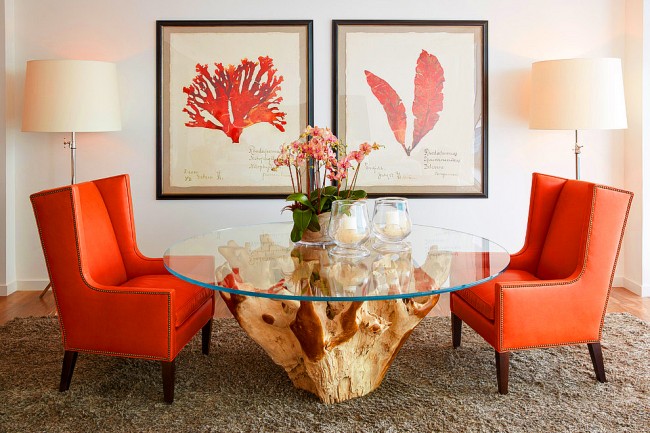 Светлые стены гостиной комнаты, оригинальный стол, строгие кресла с кораллового цвета обивкой и тематические мелочи в виде картин, цветов, свечей